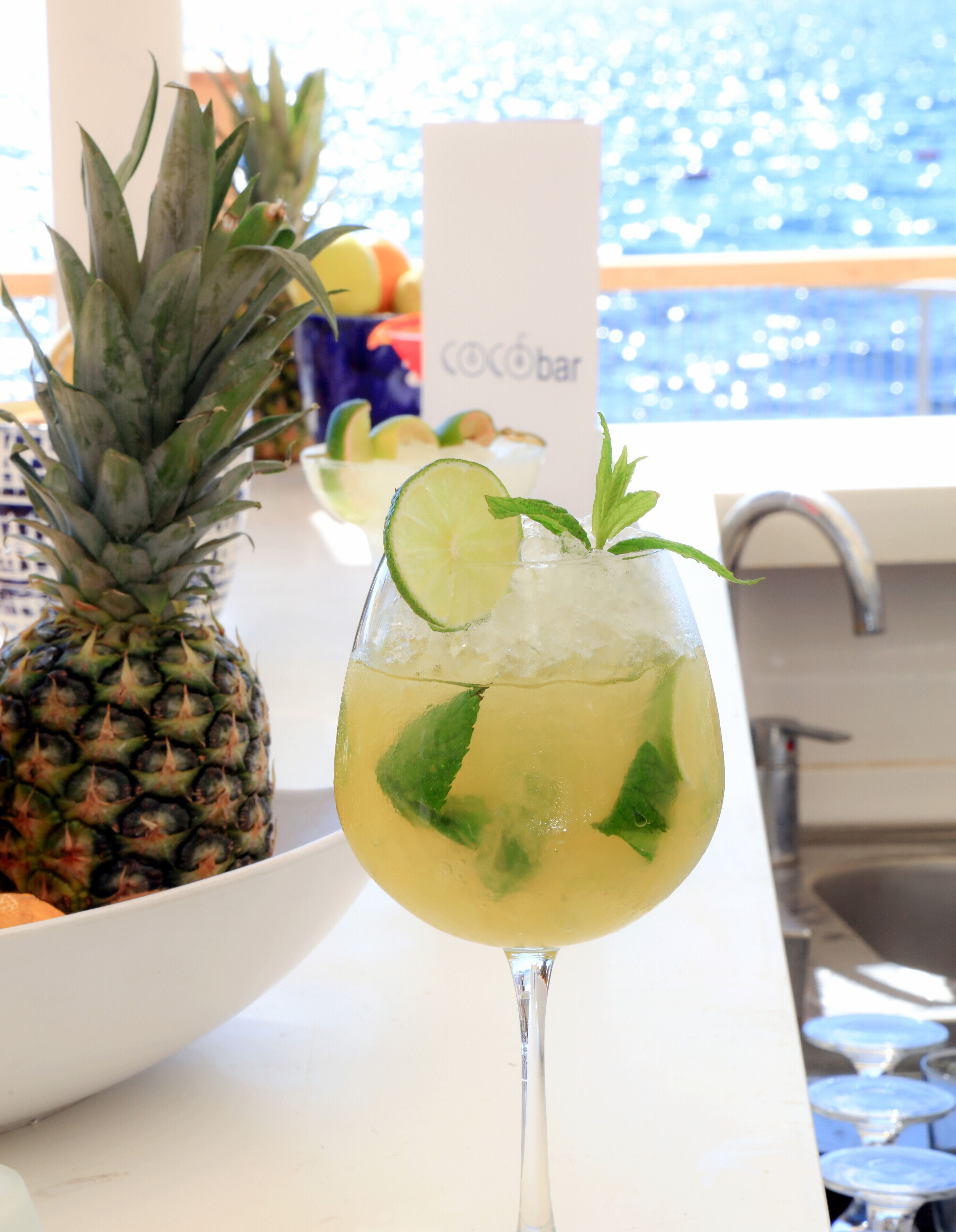 A cocktail glass with lime and mint garnish, a pineapple, and a sign reading "Cocobar" in the background.