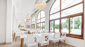 Un vaste espace dédié à la salle à manger, avec des tables et des chaises blanches, de grandes fenêtres en arc de cercle et des luminaires suspendus.