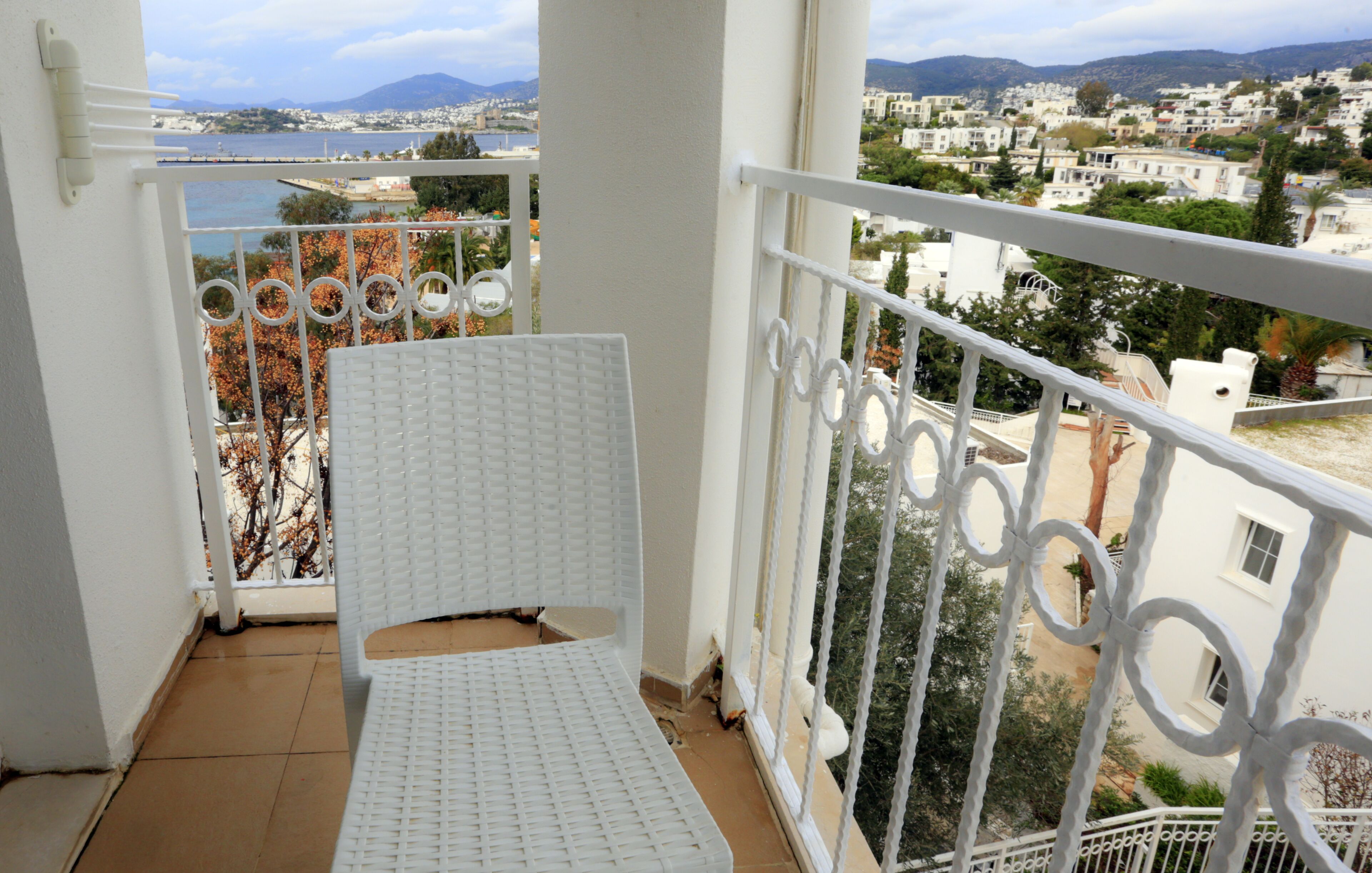 A balcony with a white metal railing, a white cushioned chair, and a view of a coastal town with buildings and trees.