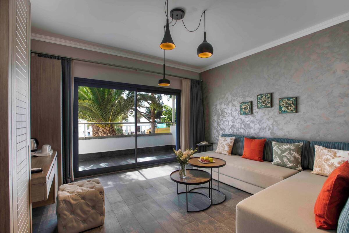 Ein modernes Wohnzimmer mit einer Couch, einem Couchtisch und einem Schreibtisch. Ein großes Fenster bietet Blick auf Palmen.