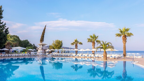 Une piscine à l’eau d’un bleu cristallin, entourée de chaises longues et de parasols, avec des palmiers et une vue sur la mer.
