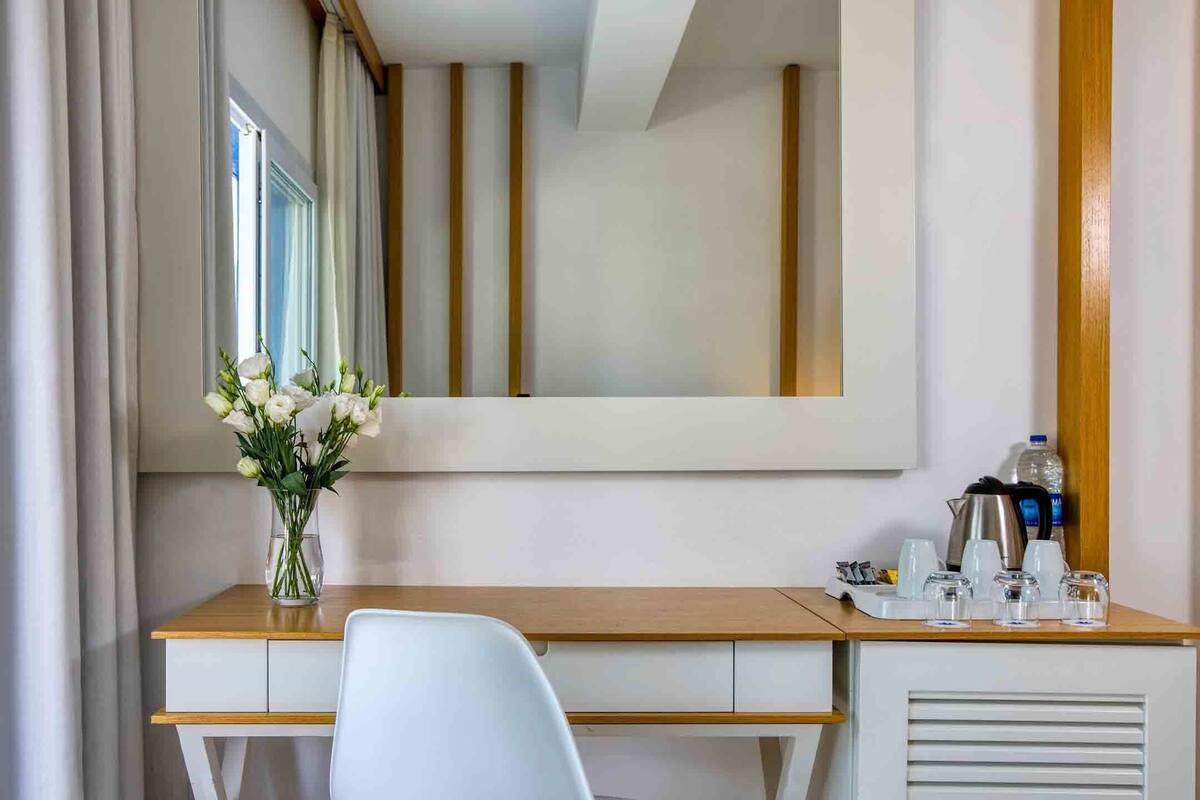 Ein Hotelzimmer mit einem Holztisch, einem weißen Stuhl, einem Spiegel, einer Vase mit Blumen und einem Tablett mit Tassen und einem Wasserkocher.