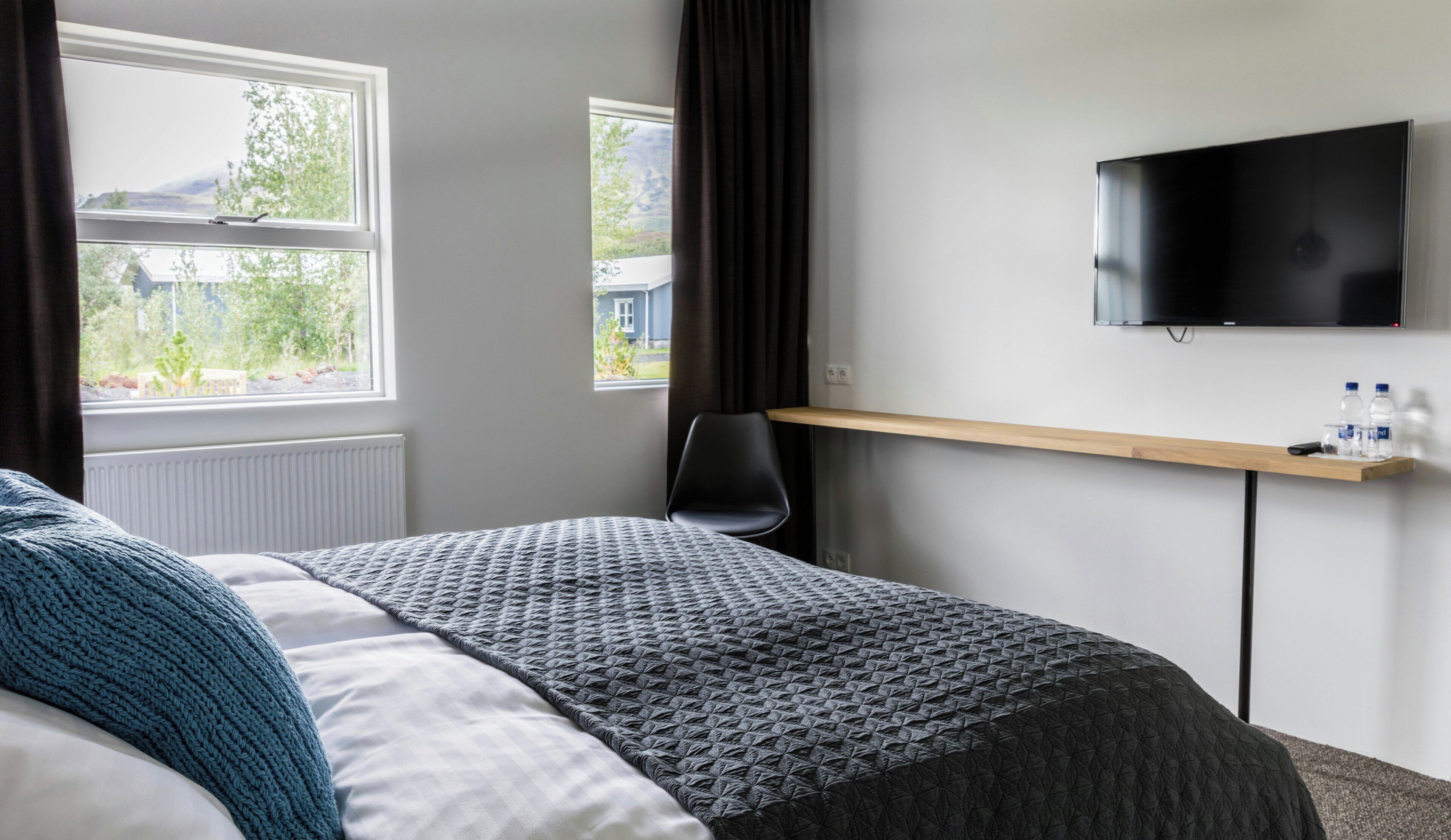 Habitación de hotel con cama, televisión montada en la pared, ventana con cortinas y estante con botellas de agua.