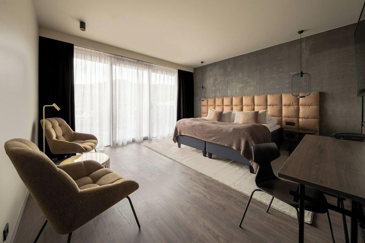 A modern hotel room with a large bed, a desk, two armchairs, and a large window with sheer curtains.