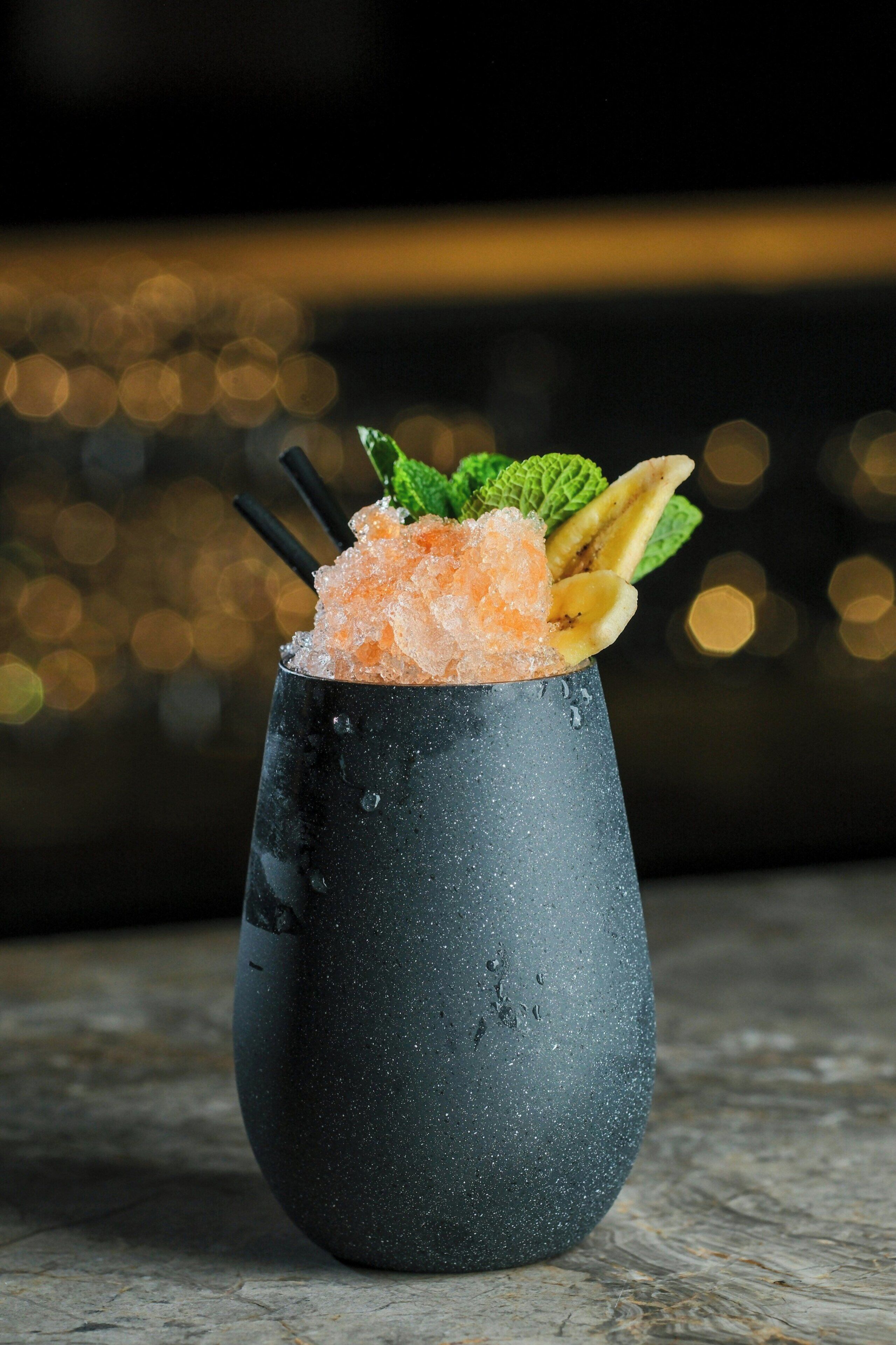 Un cóctel en un vaso texturizado, con hielo, adornado con hojas de menta y rodajas de plátano.
