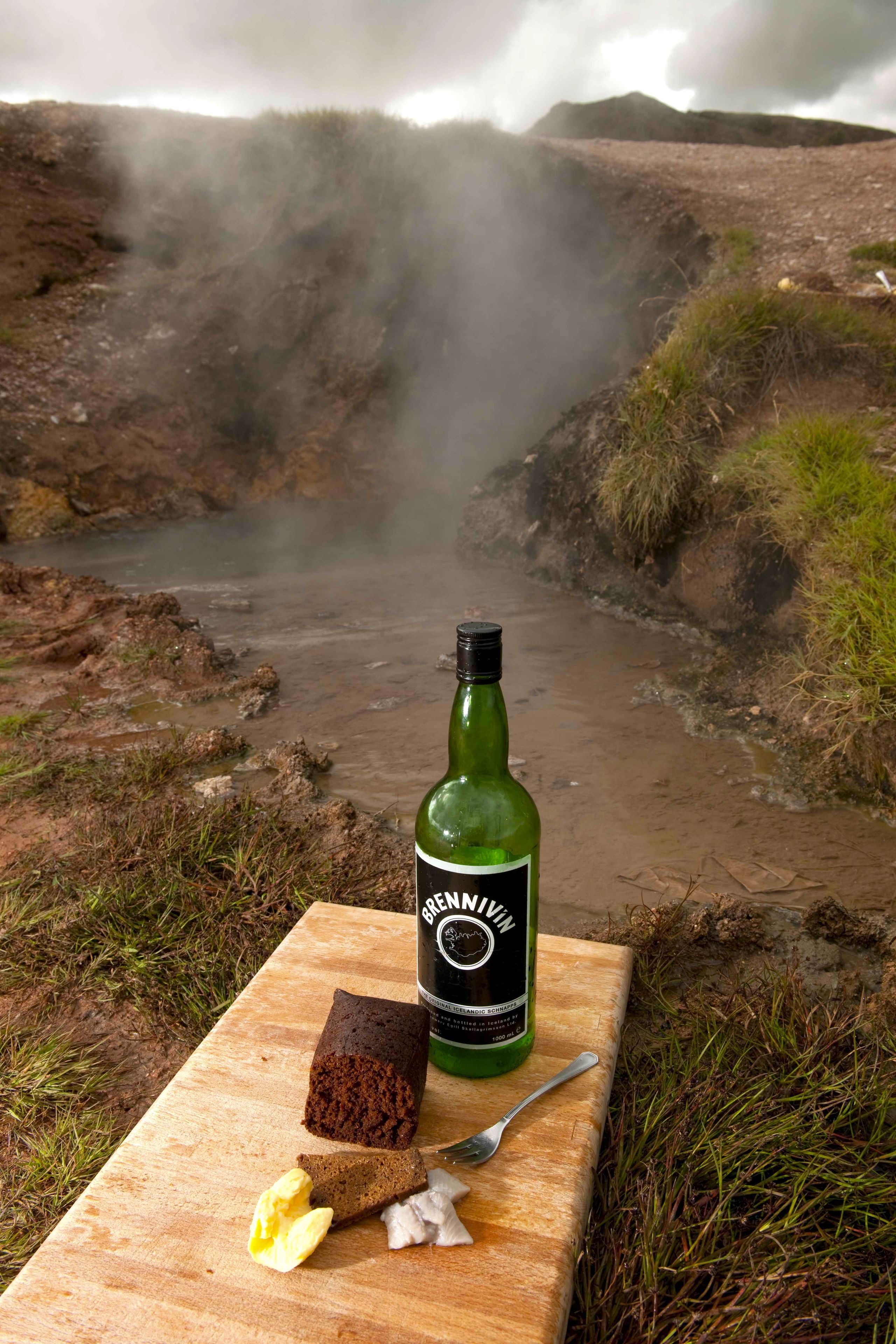 A bottle of Brennivin beer, a piece of chocolate, and a fork on a wooden board with a scenic view of a hot spring and hills in the background.