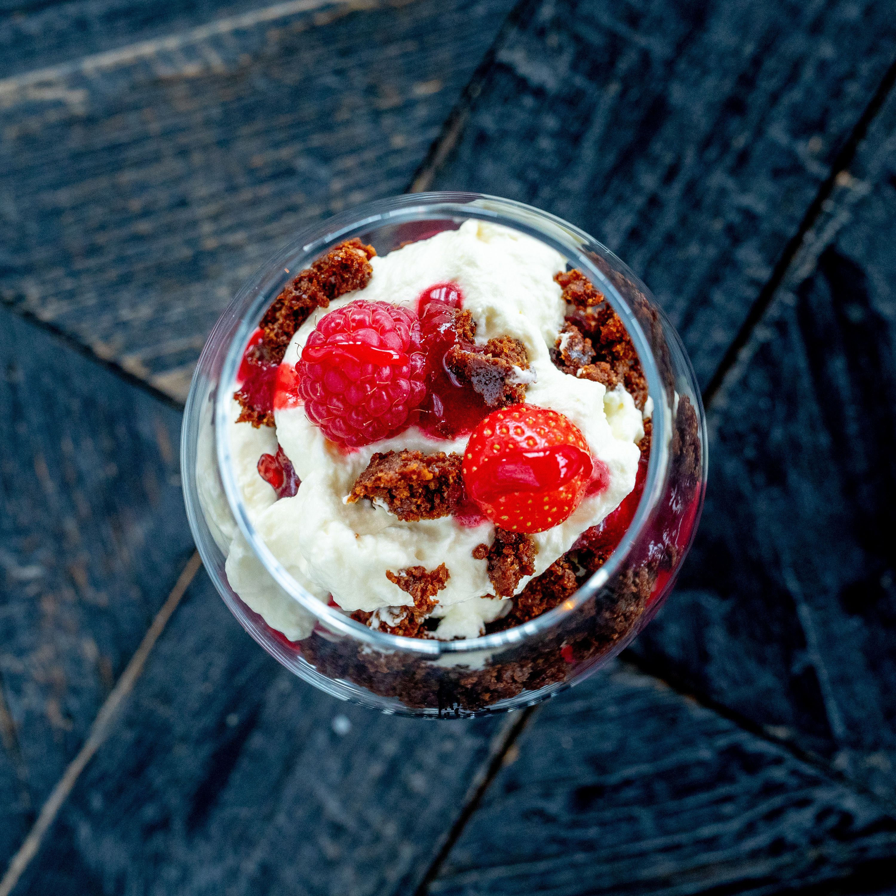 A dessert in a clear glass bowl with raspberries and a strawberry on top, placed on a dark wooden surface.