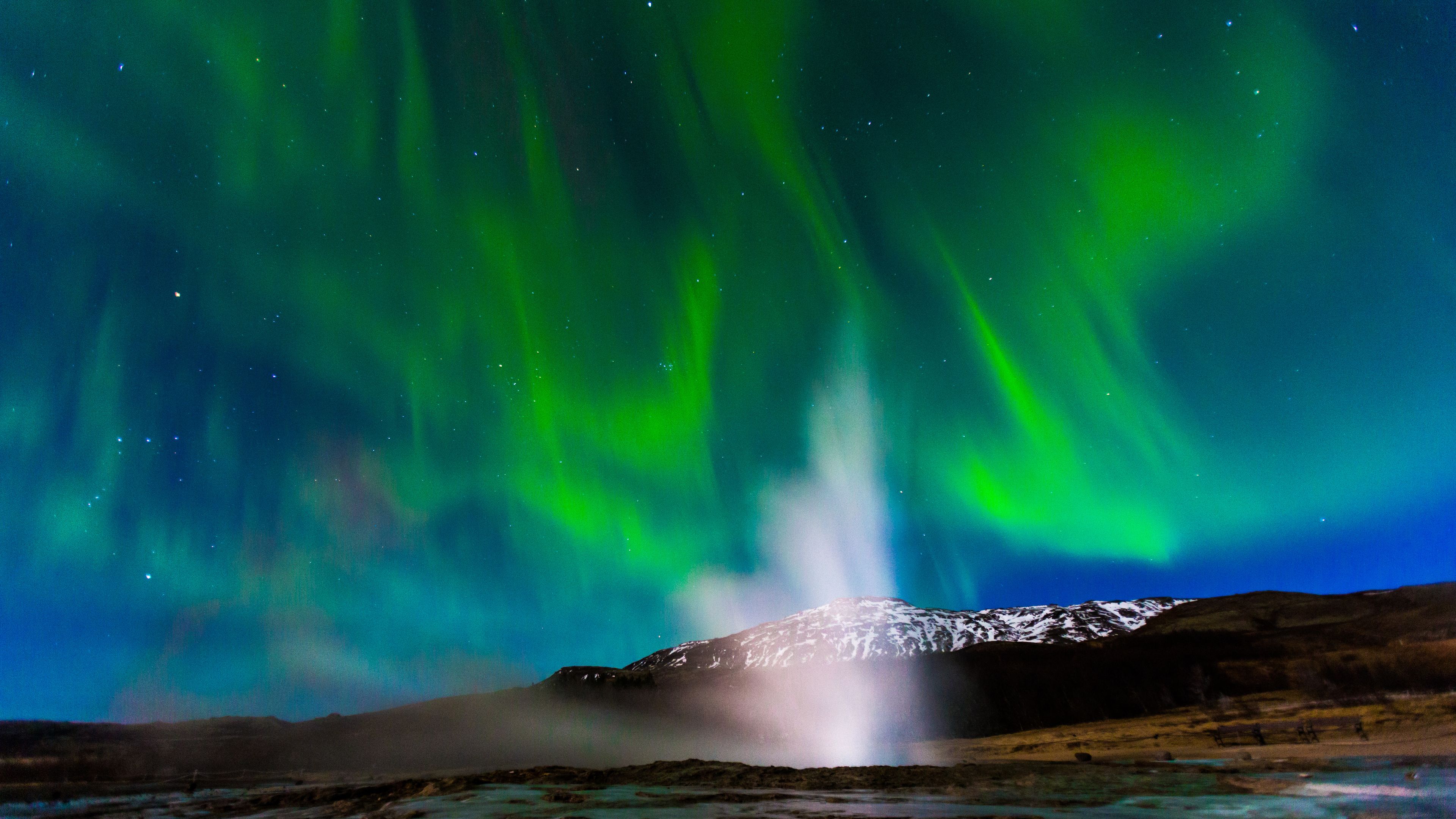 Un géiser con una montaña nevada al fondo y la aurora boreal en el cielo.