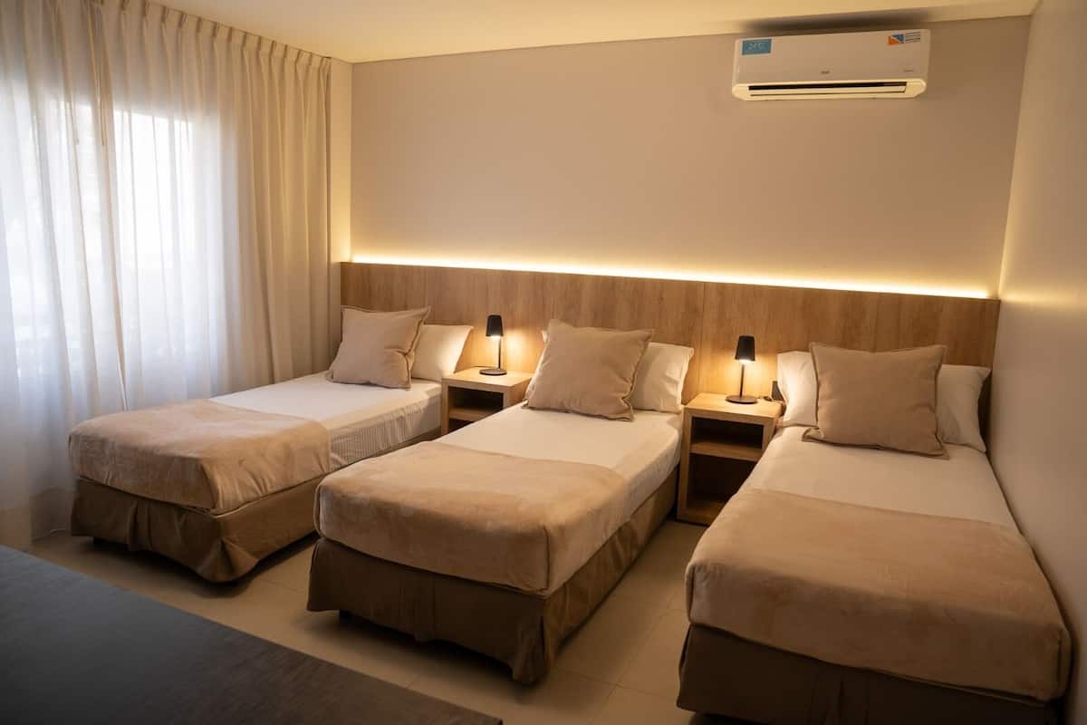 A hotel room with three single beds, a bedside table with a lamp, and an air conditioning unit on the wall.