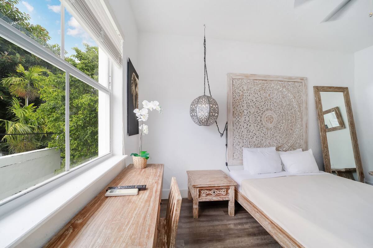 A bedroom with a bed, a bedside table, a small wooden stool, a hanging lantern, and a window with a view of greenery.