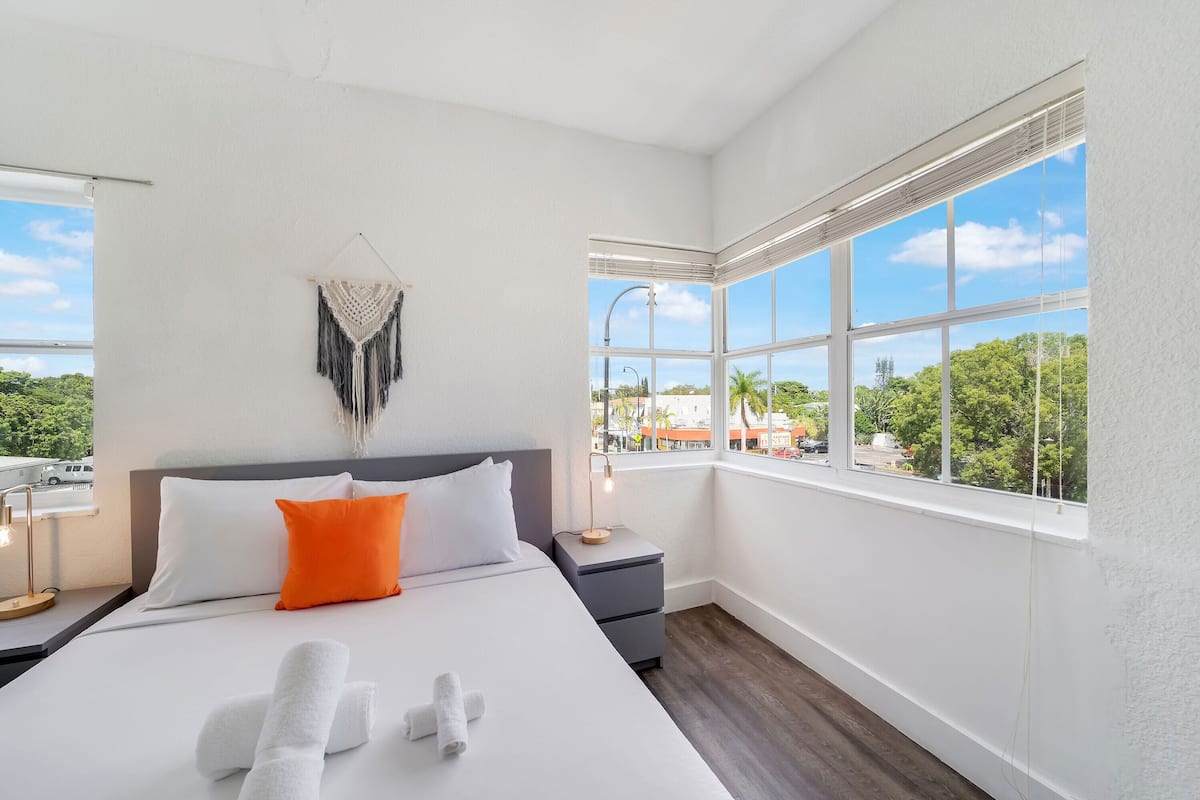 A hotel room with a large bed, a nightstand, a wall-mounted macrame piece, and a window with a view of greenery and buildings.