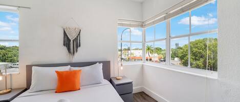 A hotel room with a large bed, a nightstand, a wall-mounted macrame piece, and a window with a view of greenery and buildings.