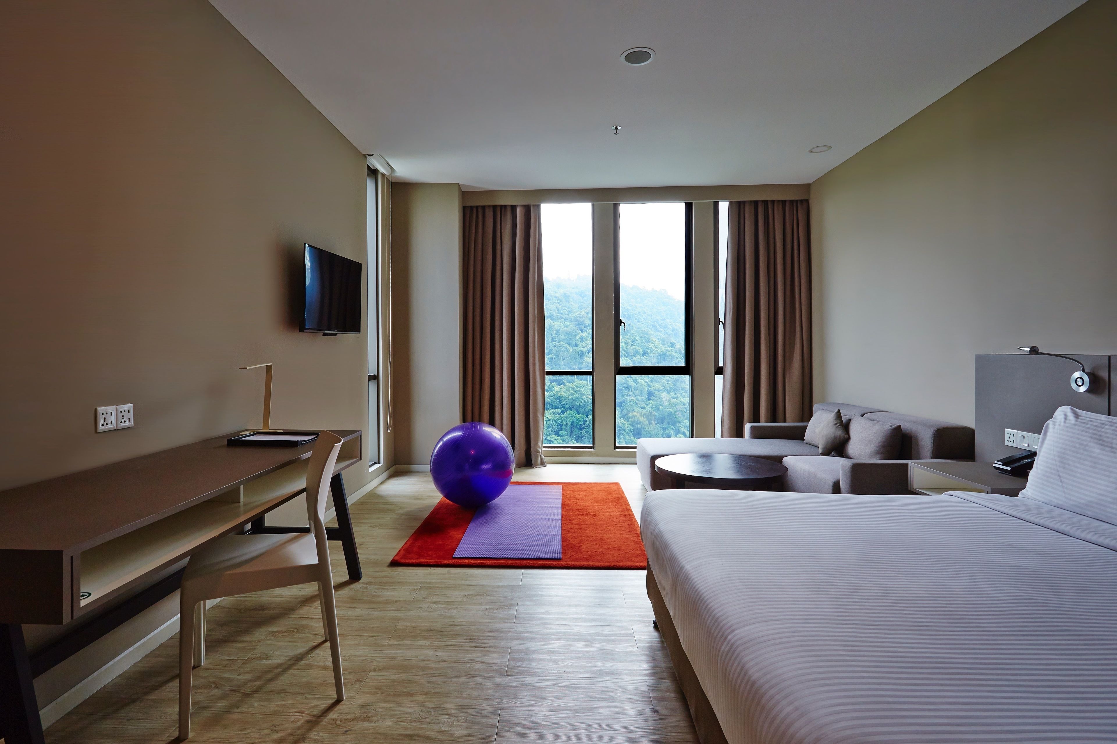 Una habitación de hotel con cama, escritorio, silla, sofá, televisión y un amplio ventanal con cortinas.