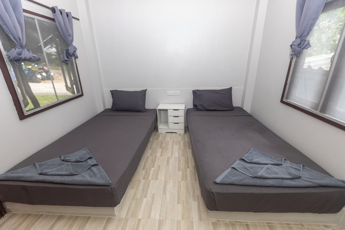 Quarto pequeno e limpo com duas camas individuais, uma mesinha de cabeceira e uma janela com cortinas.
