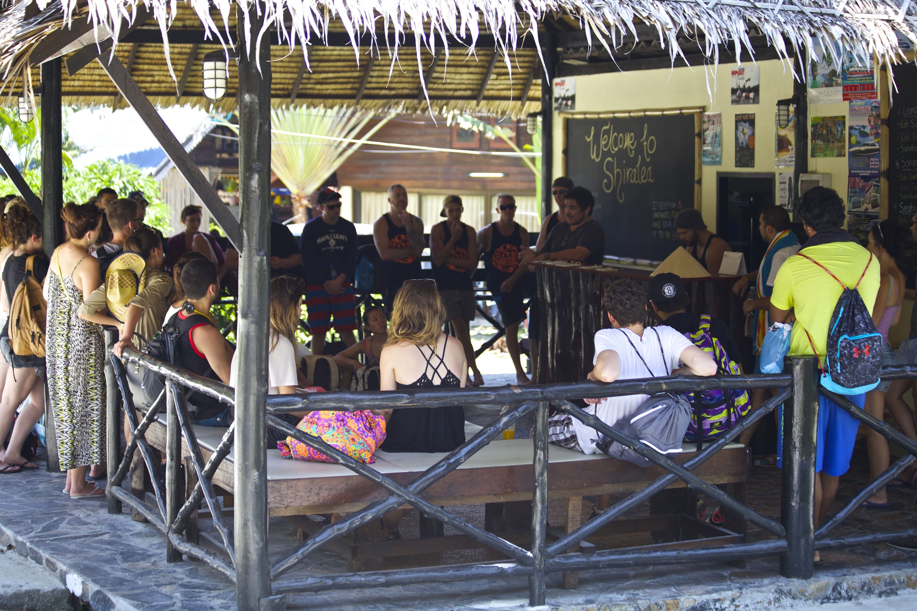 A covered outdoor seating area with a thatched roof, wooden tables, and a chalkboard sign.
