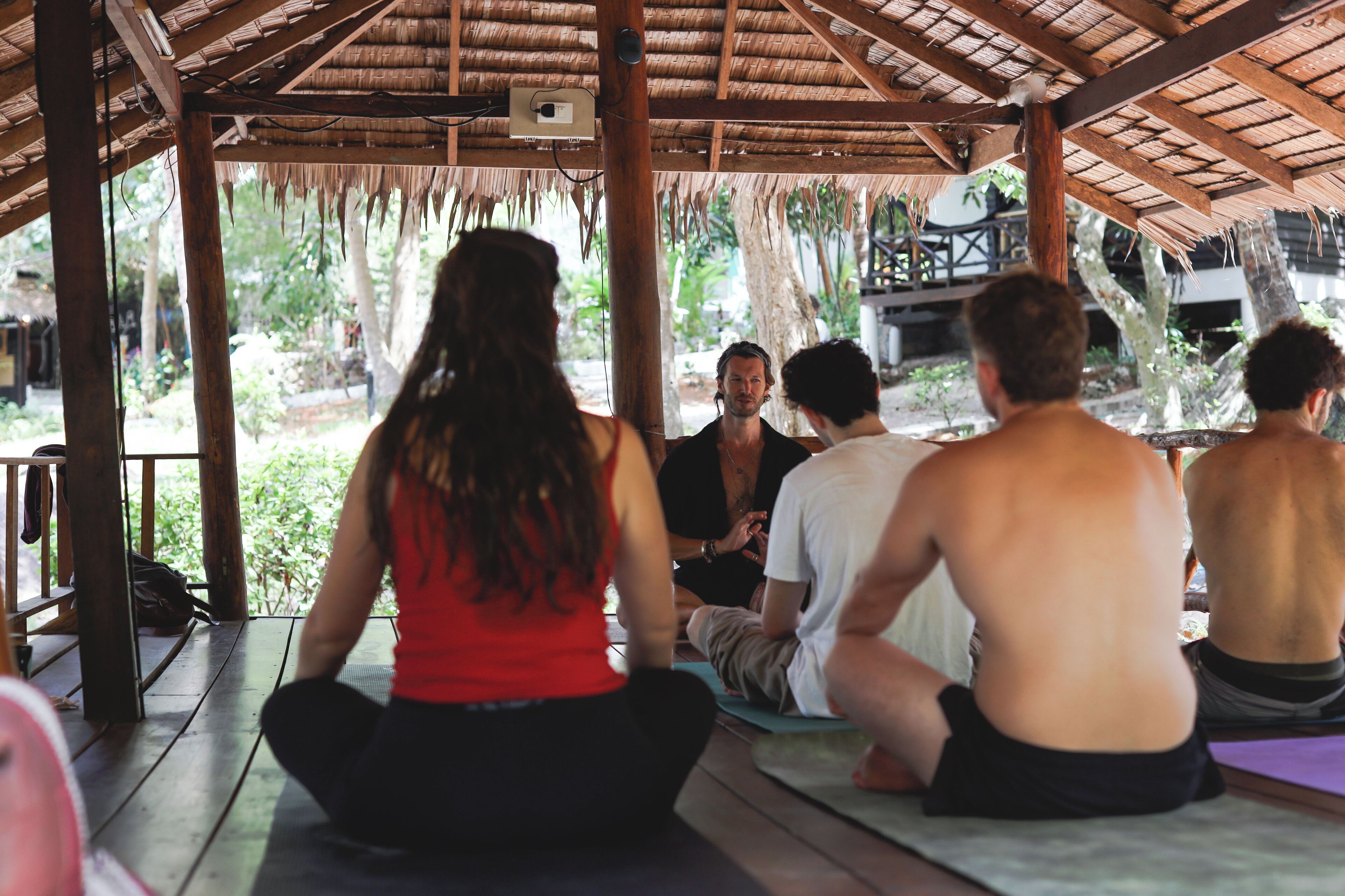 Un grupo de personas sentadas sobre esterillas de yoga en un espacio al aire libre cubierto con vigas de madera y techo de paja.