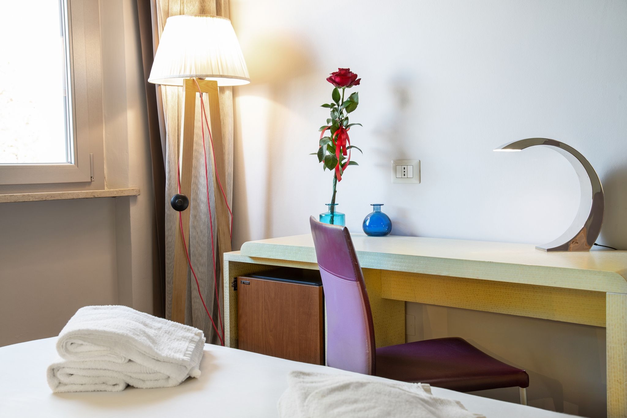 A hotel room with a bed, a desk, a chair, a lamp, a vase with a rose, and a wall socket.