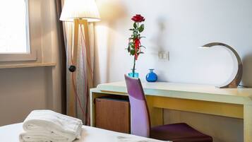 A hotel room with a bed, a desk, a chair, a lamp, a vase with a rose, and a wall socket.