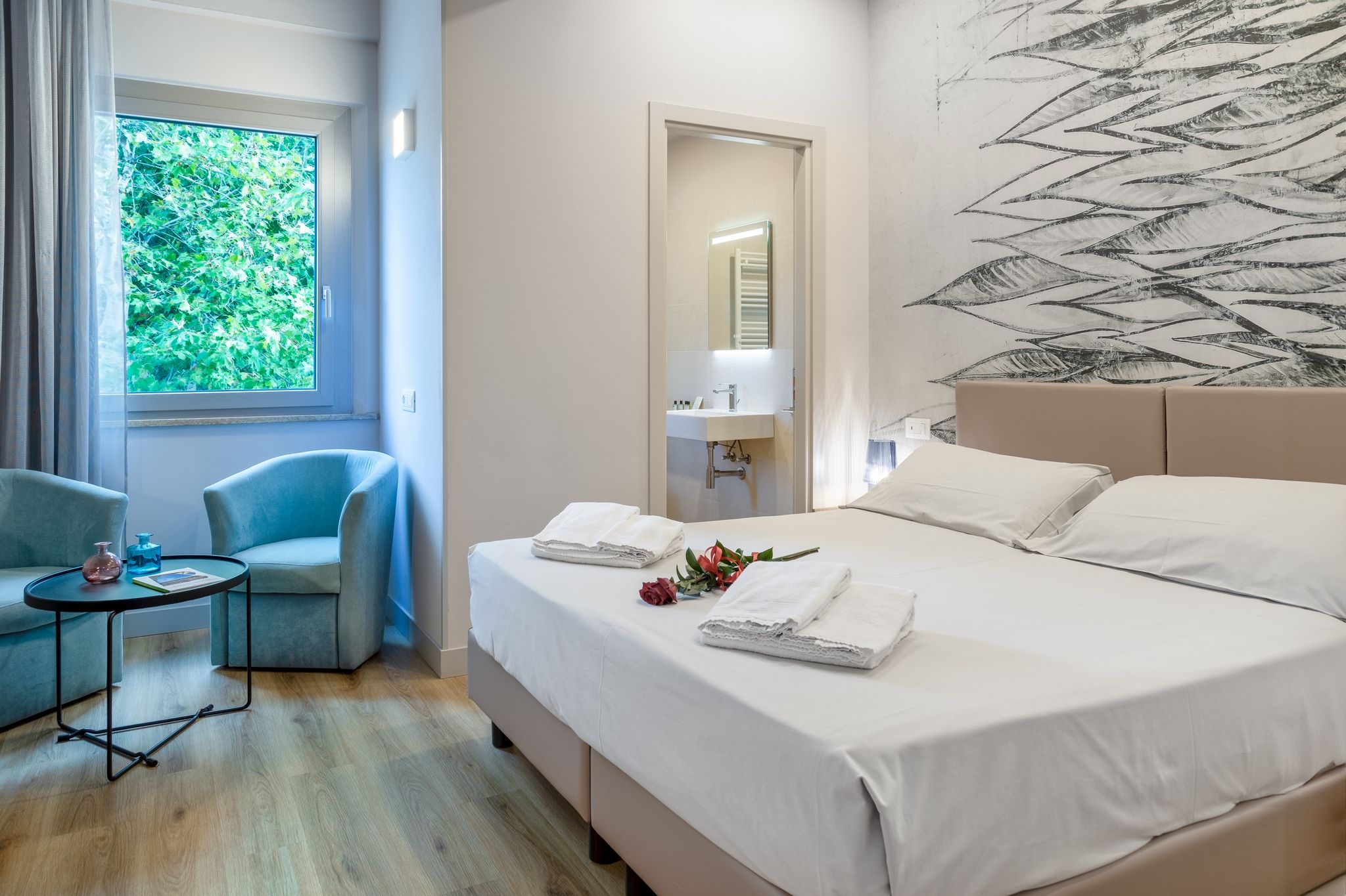 Habitación de hotel con cama, una silla, una mesita, ventana con vistas a vegetación y pared con diseño abstracto blanco y negro.