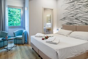 Habitación de hotel con cama, una silla, una mesita, ventana con vistas a vegetación y pared con diseño abstracto blanco y negro.