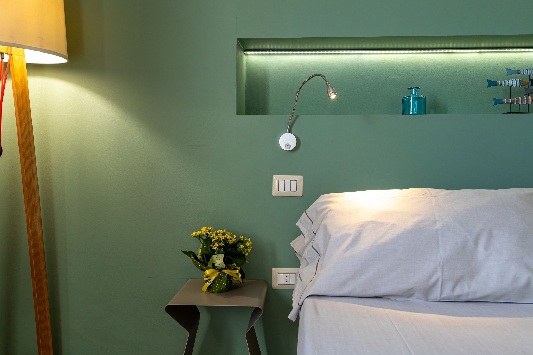 Quarto com cama, mesa de cabeceira com um vaso de flores, luminária, prateleira na parede com artigos de higiene e uma tomada.