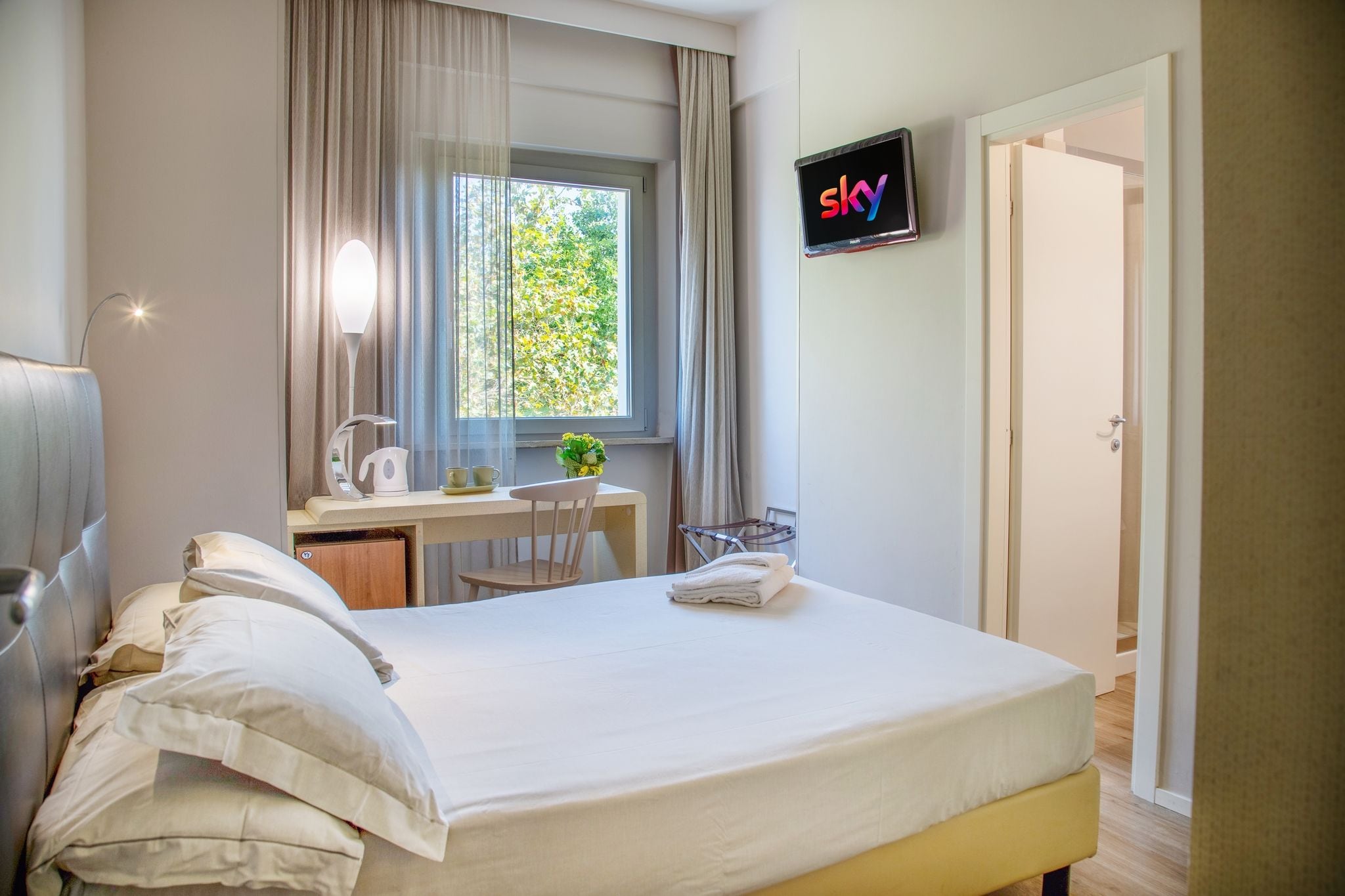 A hotel room with a bed, a desk with a chair, a TV mounted on the wall, and a window with curtains.