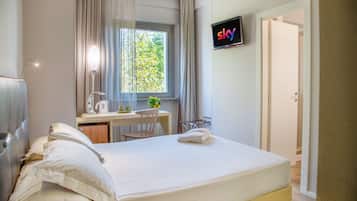 A hotel room with a bed, a desk with a chair, a TV mounted on the wall, and a window with curtains.