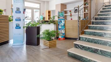 A hotel lobby with wooden flooring, a staircase with marble steps, and various plants and promotional posters.