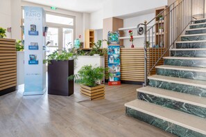 A hotel lobby with wooden flooring, a staircase with marble steps, and various plants and promotional posters. - Hotel De La Ville (Fano)