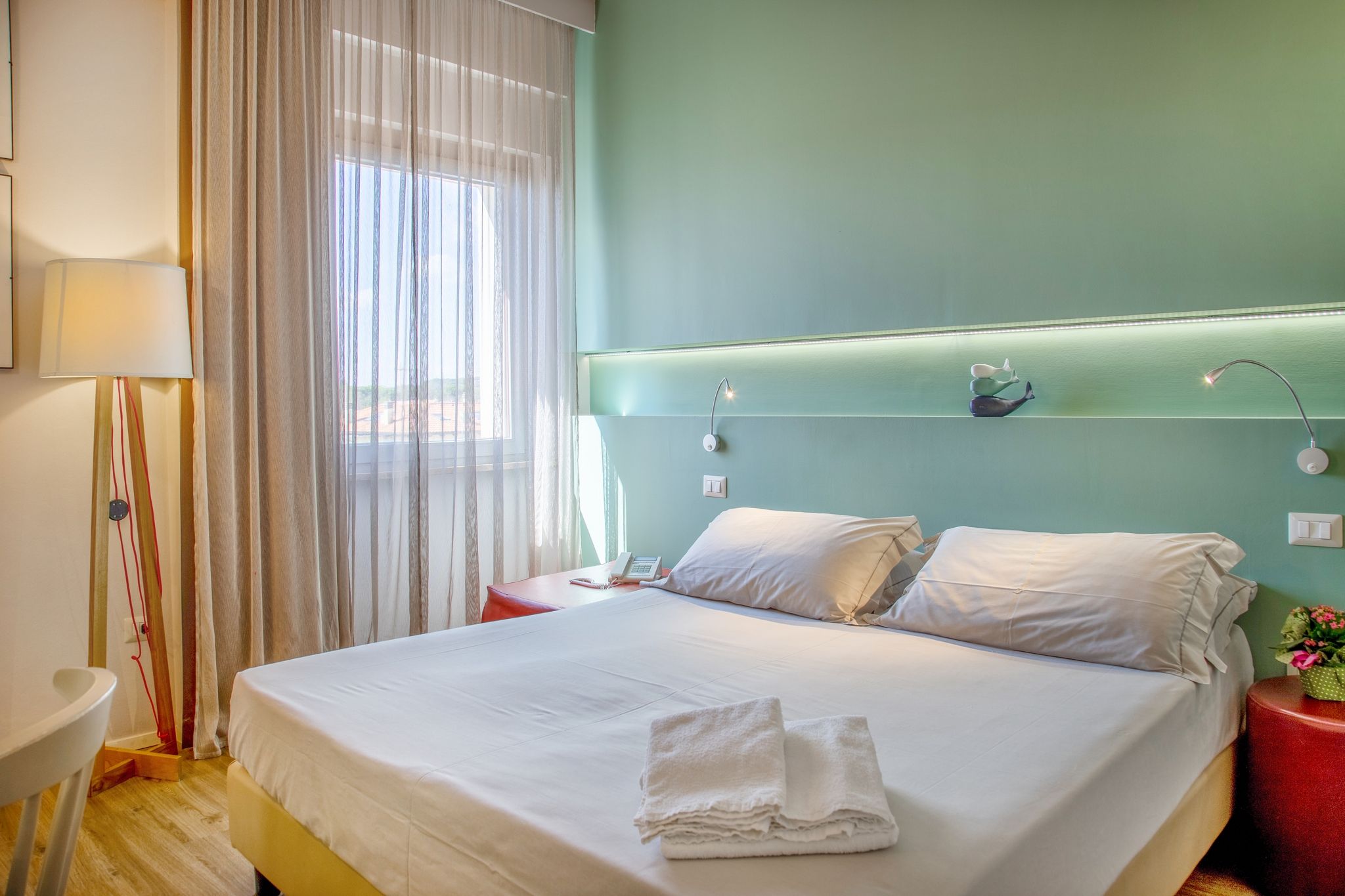 Une chambre d’hôtel avec un lit, une table de chevet, une lampe et une fenêtre avec des rideaux légers.