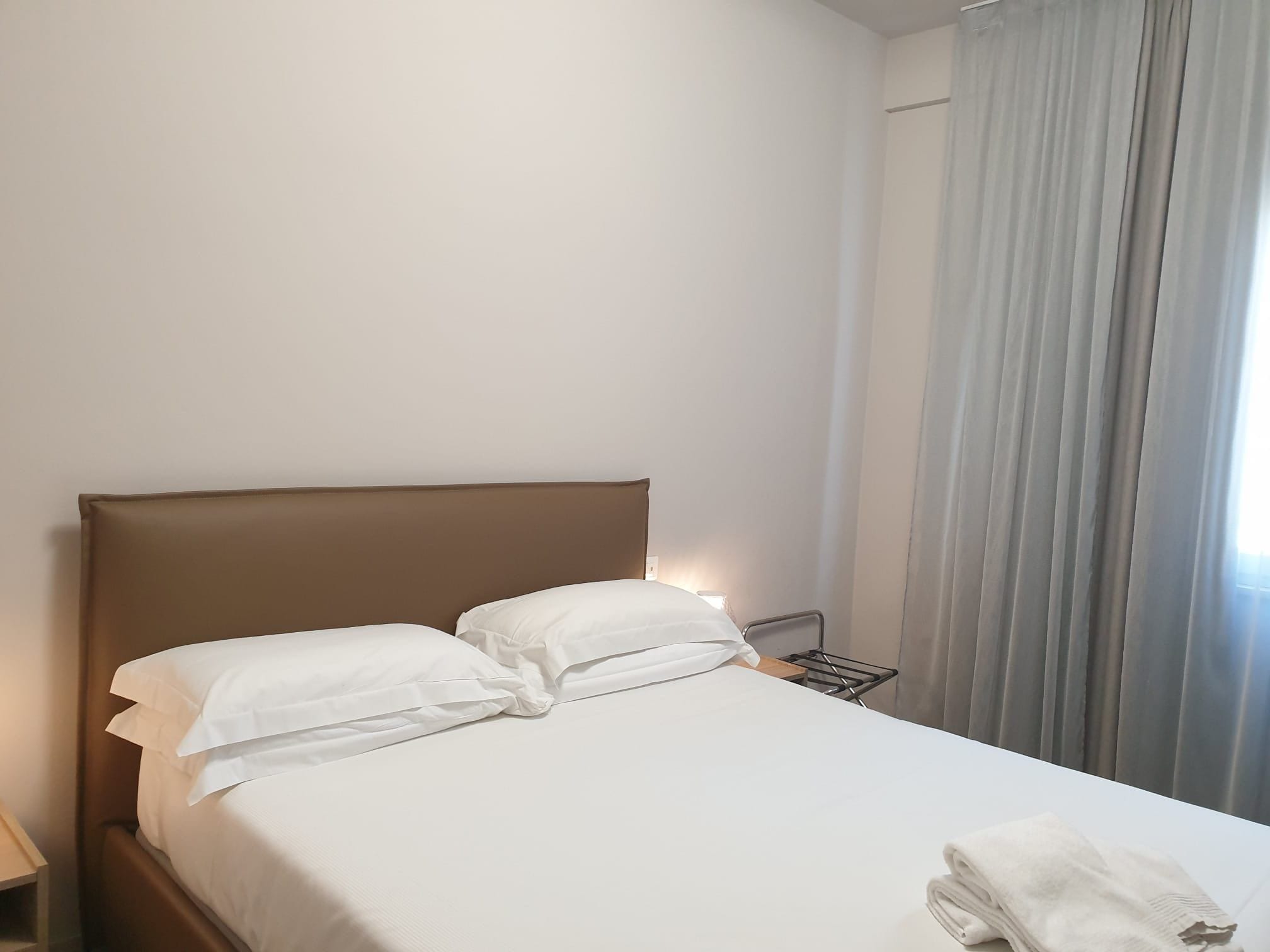 A hotel room with a bed, pillows, a nightstand, a lamp, and a window with curtains.