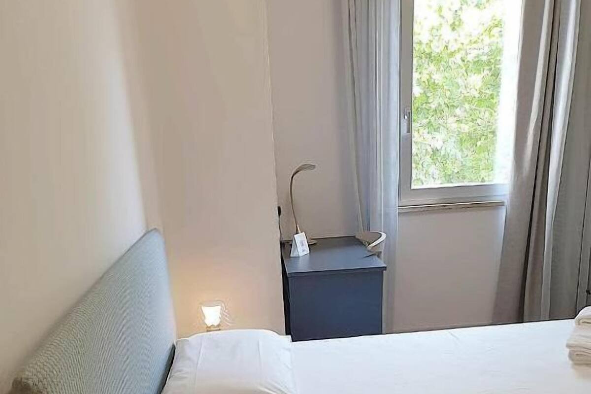 A neatly made bed in a hotel room with a headboard, a bedside table with a lamp, and a window with curtains.