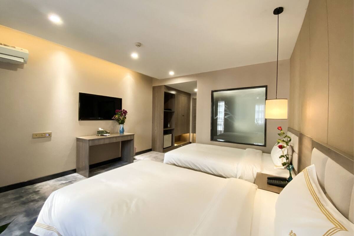 A hotel room with two beds, a bedside table with a lamp, a mirror, a desk with a vase of flowers, and a large window.