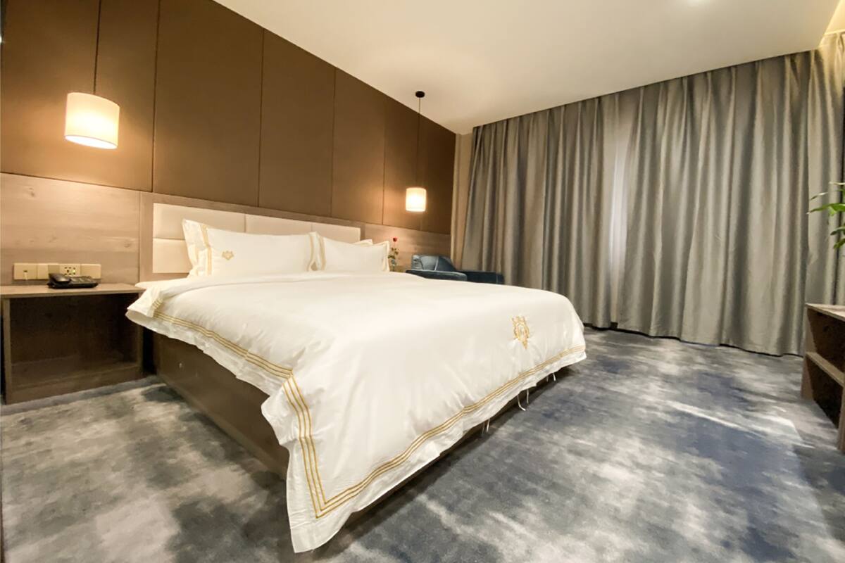 A large bed with a white bedspread and gold trim, a nightstand with a phone, and a window with curtains.