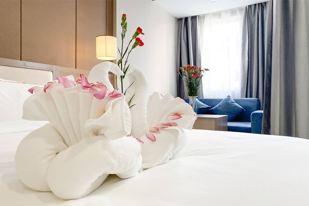 A neatly made bed with a decorative swan-shaped towel arrangement, a bedside lamp, a vase with flowers, and a blue sofa in a hotel room.
