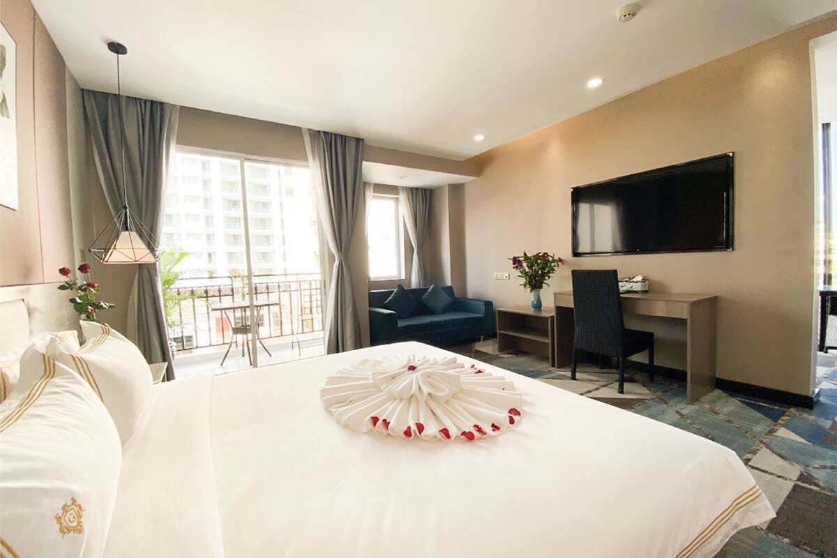 A hotel room with a large bed, a desk, a chair, a TV, and a balcony with a view of buildings.