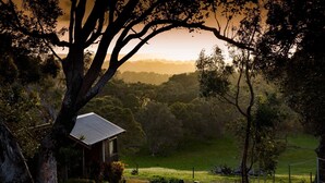A rustic house with a sloped roof, surrounded by a fenced property with a grassy field and dense trees in the background. - Wildwood Valley (Yallingup)