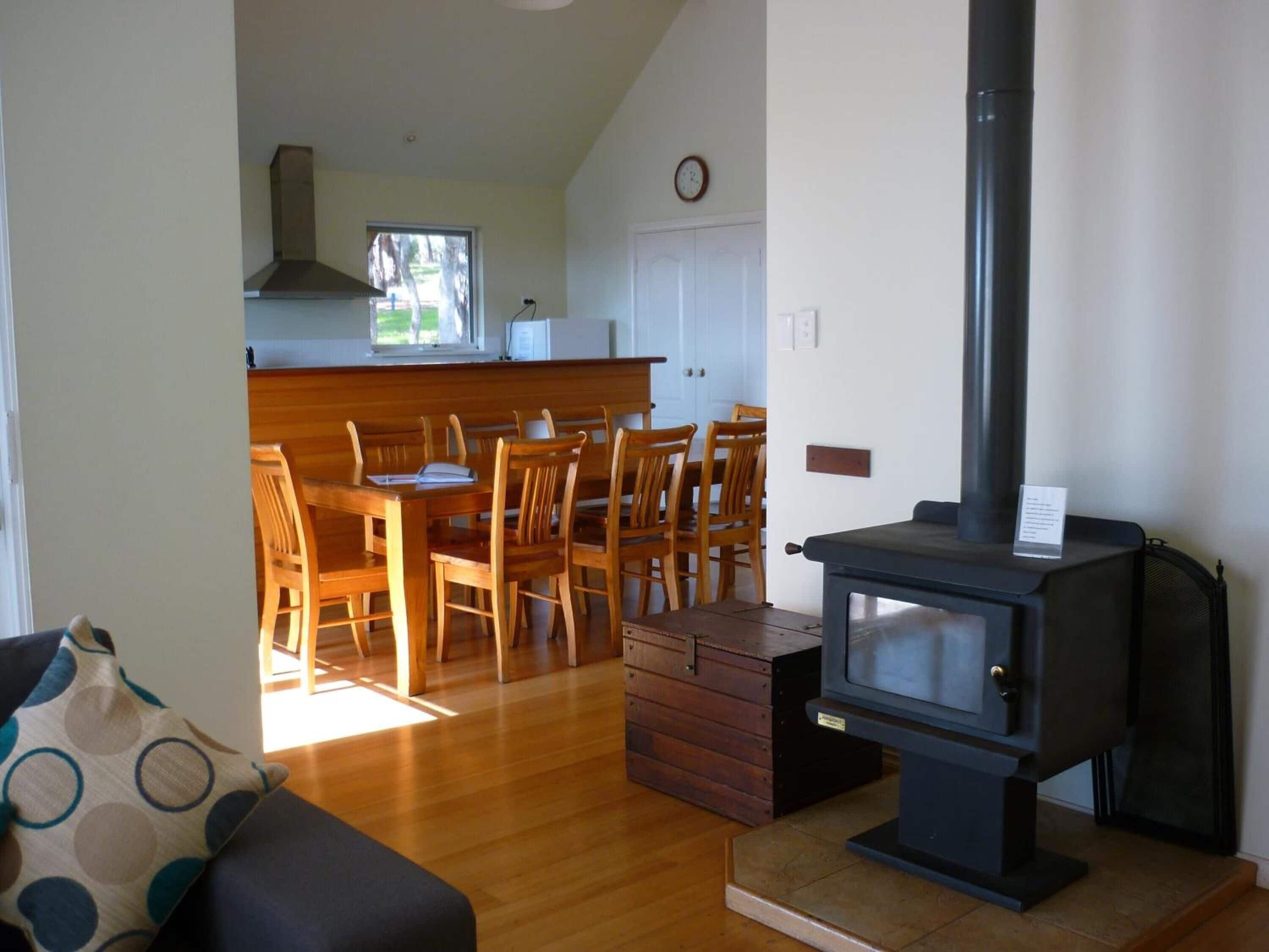 A cozy living room with a wooden dining table and chairs, a black wood-burning stove, and a window providing natural light.