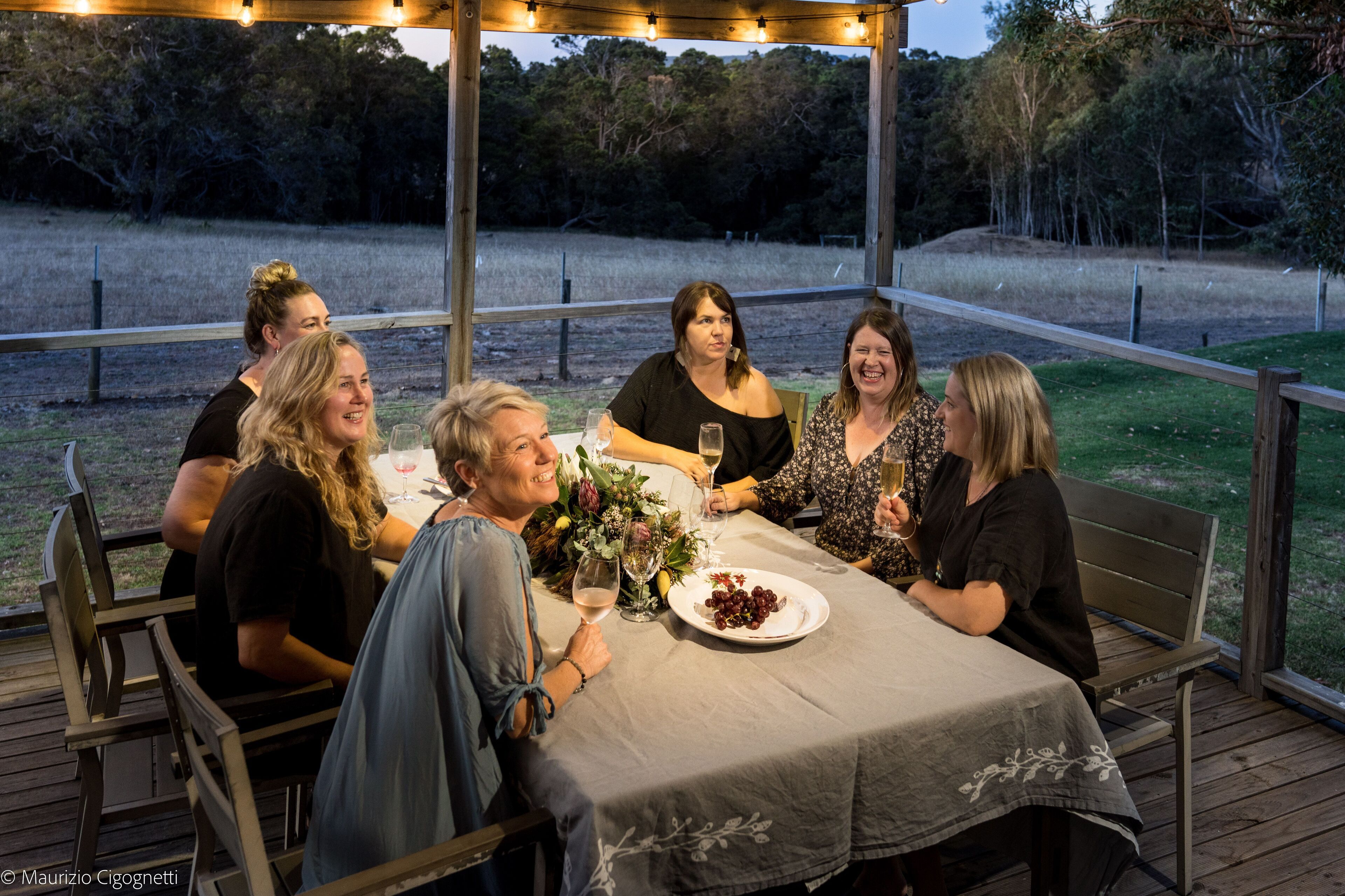 A group of people seated at an outdoor table with a tablecloth, surrounded by wooden chairs and benches, with a view of a grassy field and trees in the background.