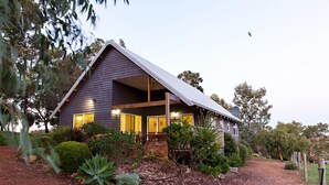 A two-story house with a dark exterior, a covered porch, and a well-maintained garden with various plants and a gravel driveway. - Wildwood Valley (Yallingup)