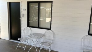 A building exterior with a closed door, a window with a metal screen, and two white metal chairs.