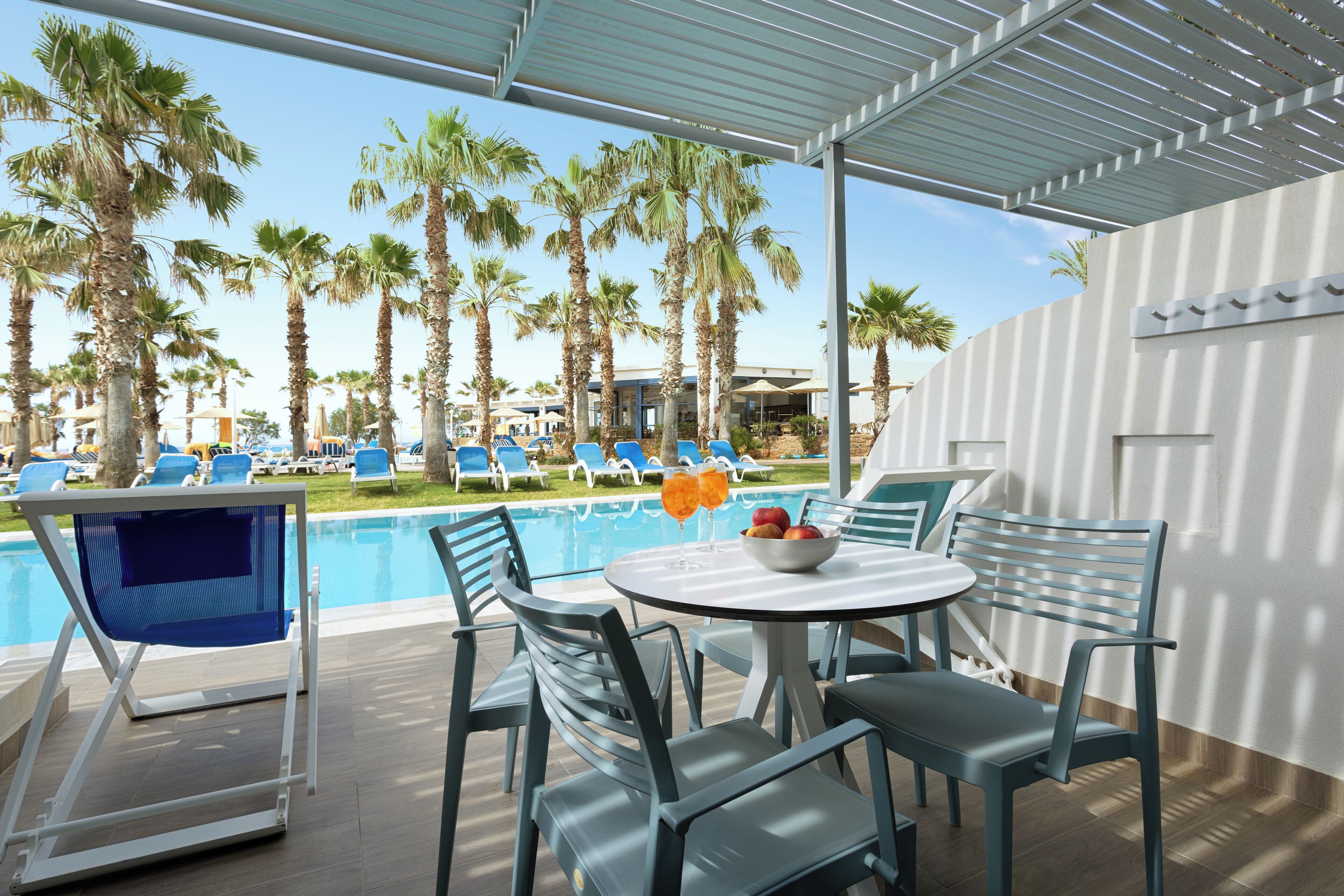 Zona junto a la piscina con sillas, una mesa, palmeras y vista despejada del agua.