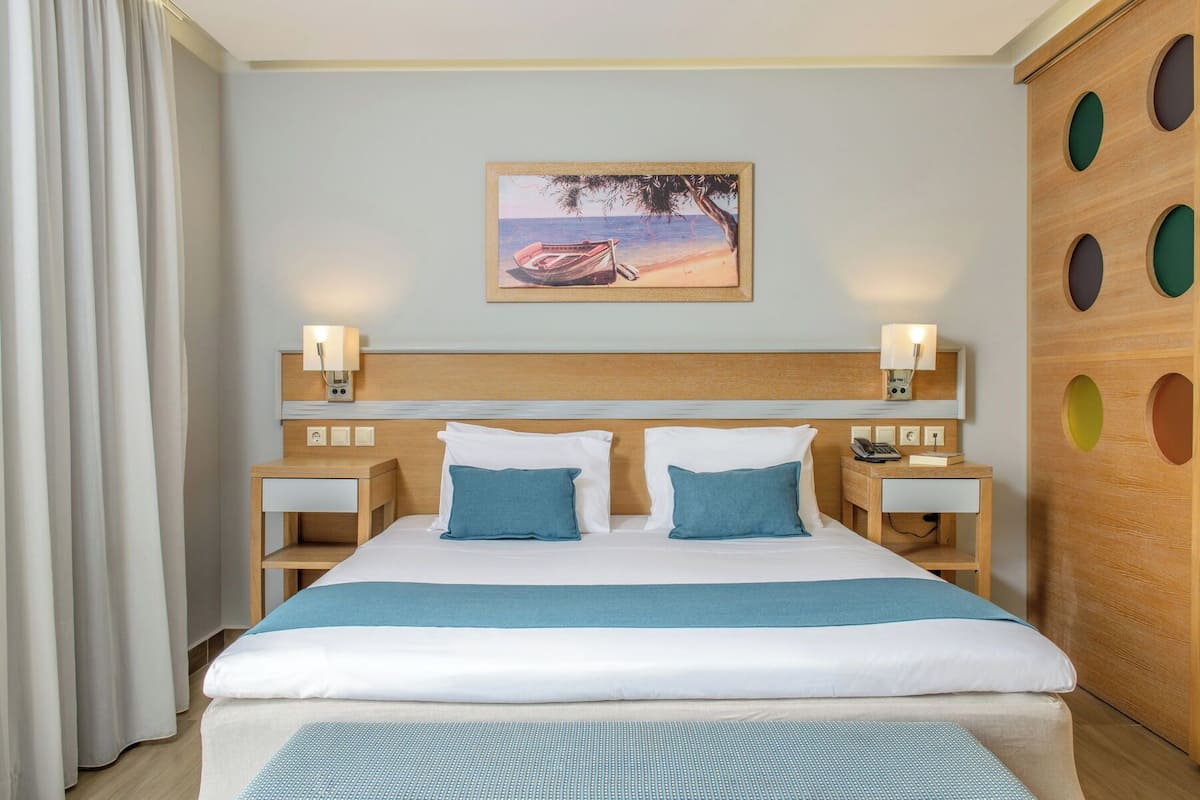 A hotel room with a bed, two bedside tables, a nightstand, a wall-mounted lamp, and a painting of a beach scene.