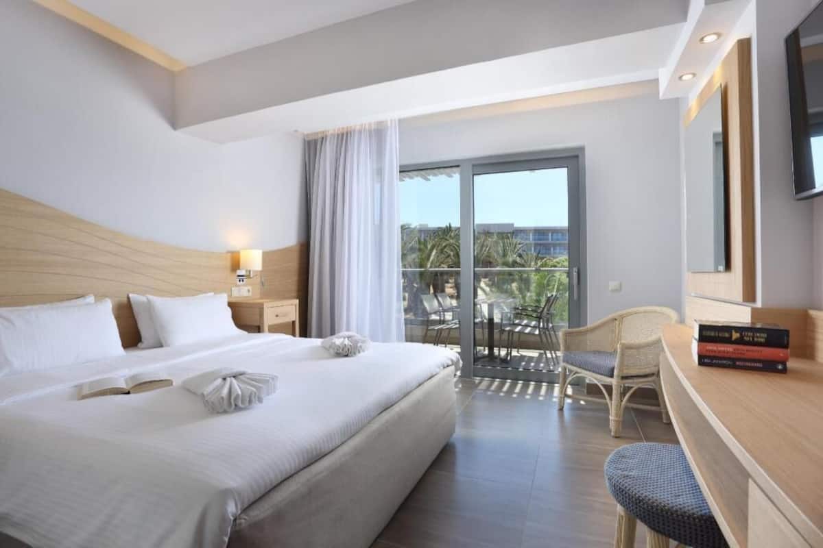 A hotel room with a large bed, a balcony with outdoor furniture, a TV, and a bookshelf.
