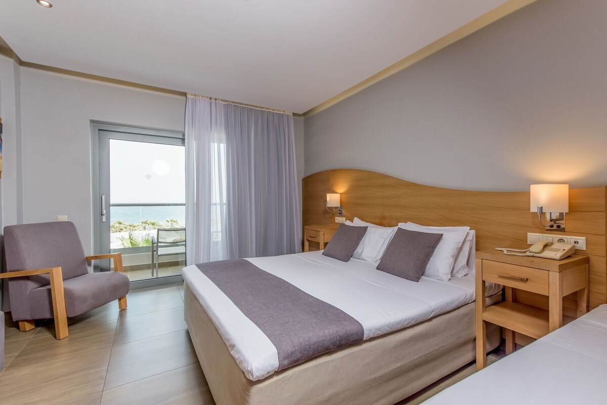 A hotel room with a bed, a chair, a bedside table with a lamp, and a view of the sea.