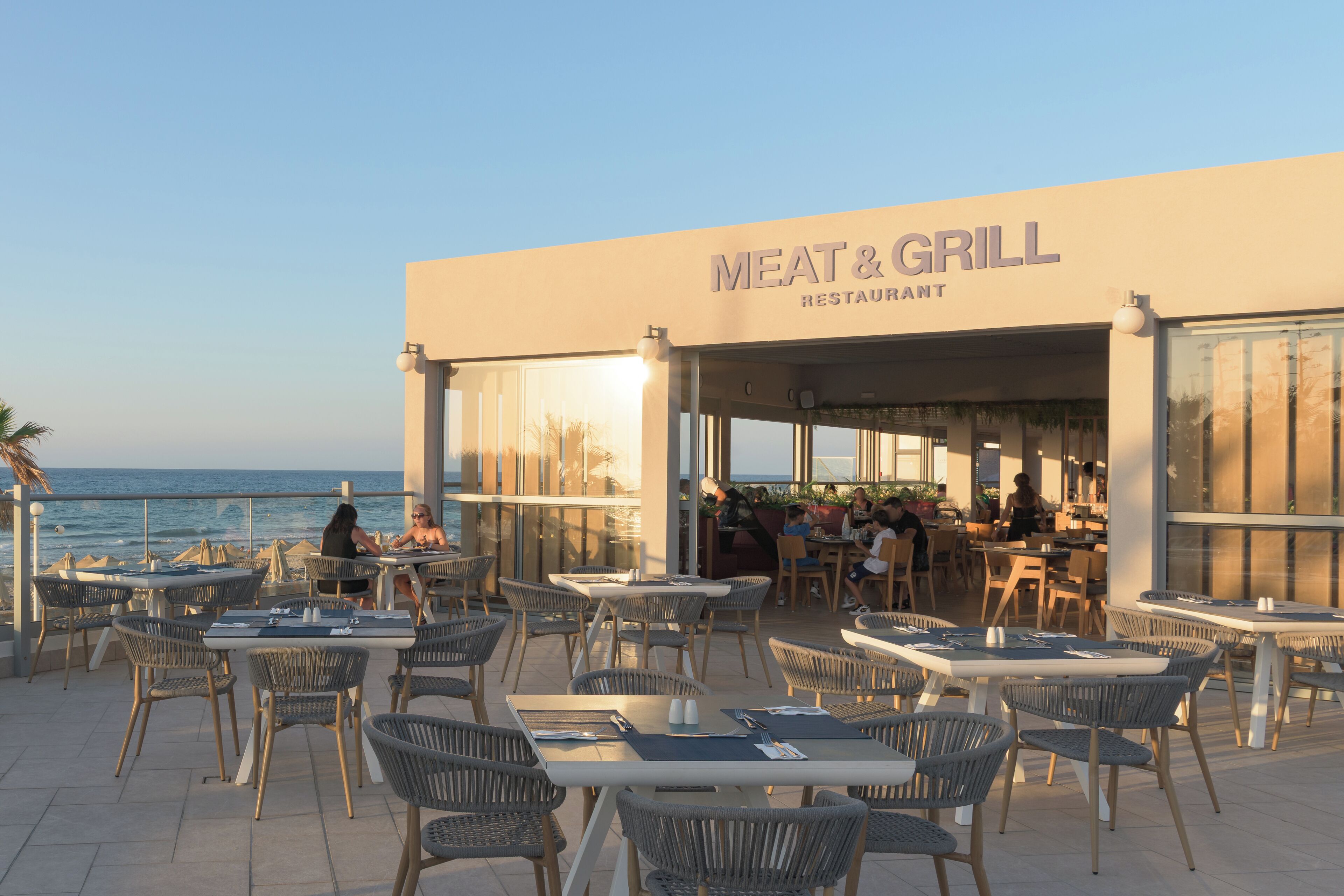 An outdoor seating area with tables and chairs, a view of the ocean, and a building labeled 'MEAT & GRILL RESTAURANT.'