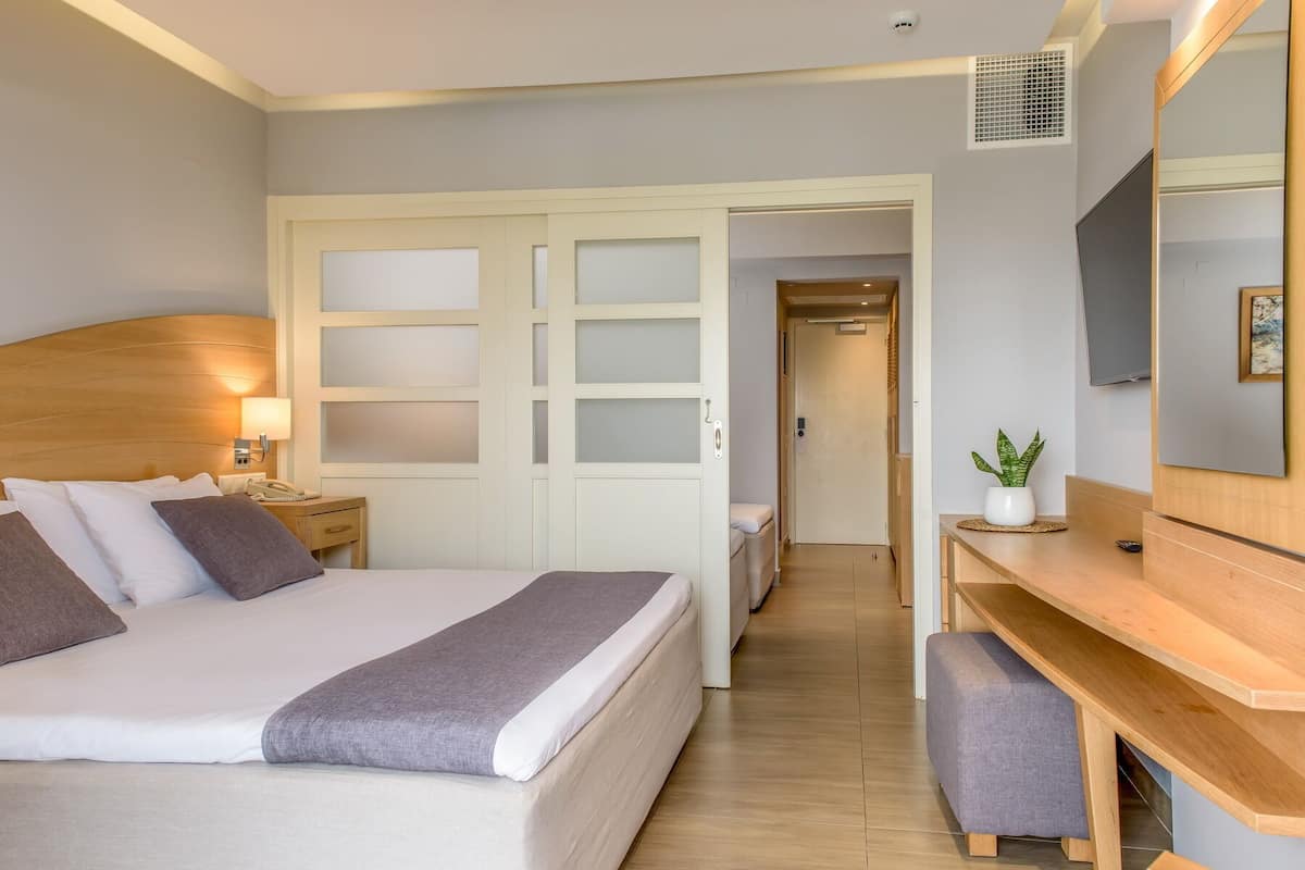 A hotel room with a bed, bedside table, lamp, wardrobe, desk, chair, potted plant, and a large mirror.