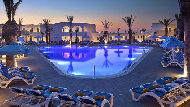 Ein Pool mit Liegestühlen und Sonnenschirmen, umgeben von Palmen und Gebäuden.