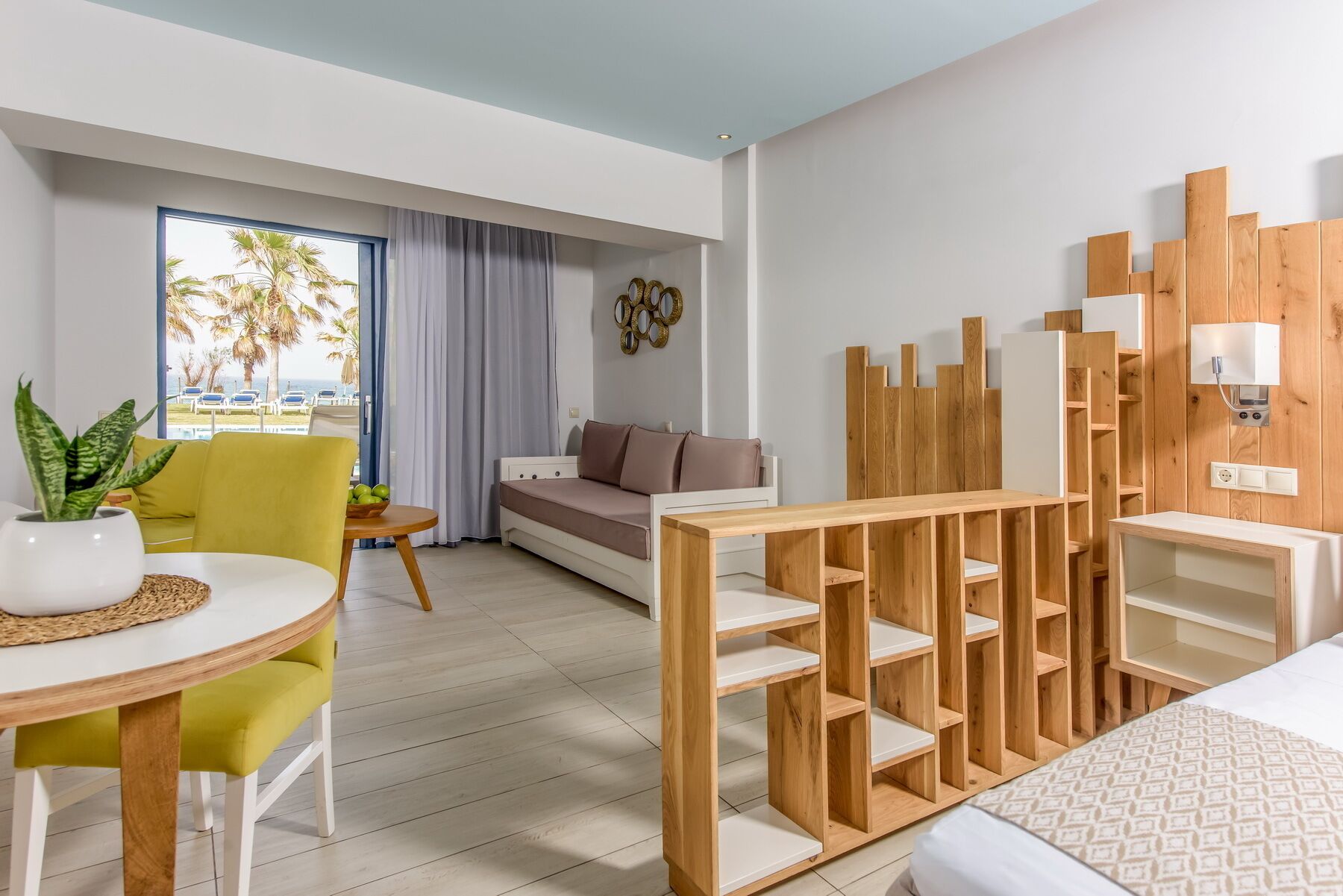 Ein modernes Hotelzimmer mit einer Couch, einem Esstisch mit Stühlen, einer Holzhülle und Blick auf Palmen und das Meer.