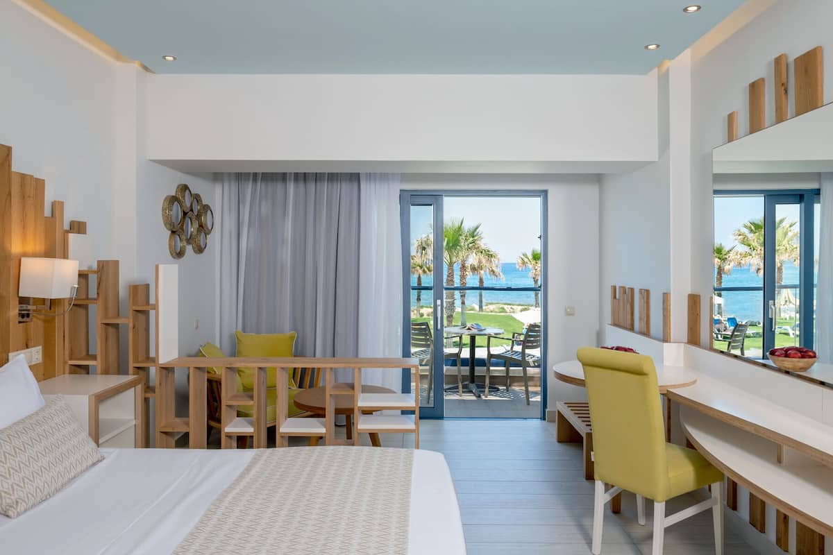A hotel room with a bed, a desk, and a dining area with chairs. There is a view of the ocean and palm trees through the window.